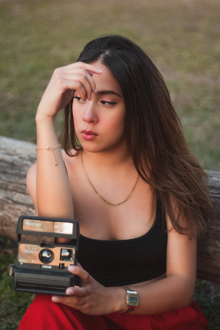 Young woman in casual attire holding a vintage camera, posing outdoors with a thoughtful expression.